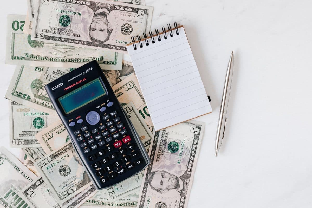 A calculator, a small notepad and a pen lying on scattered US dollar bills on a white table