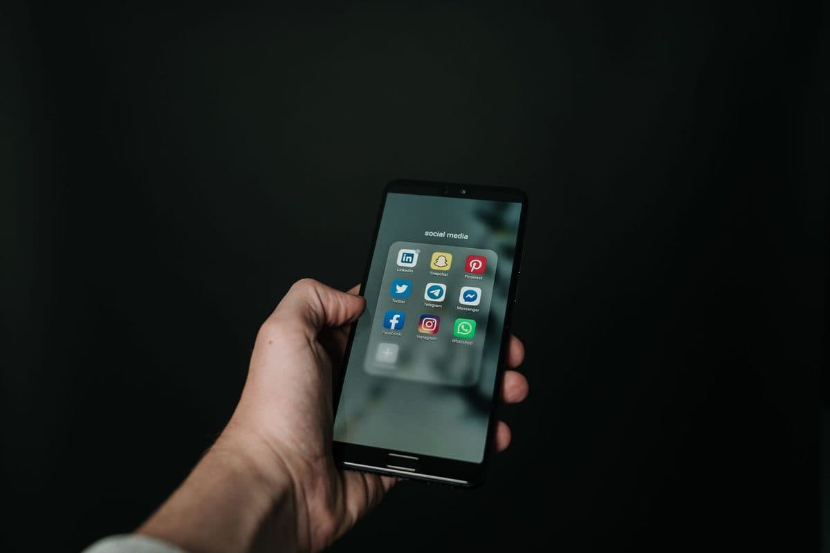 A hand holding a smartphone showing a folder of social media apps against a dark background
