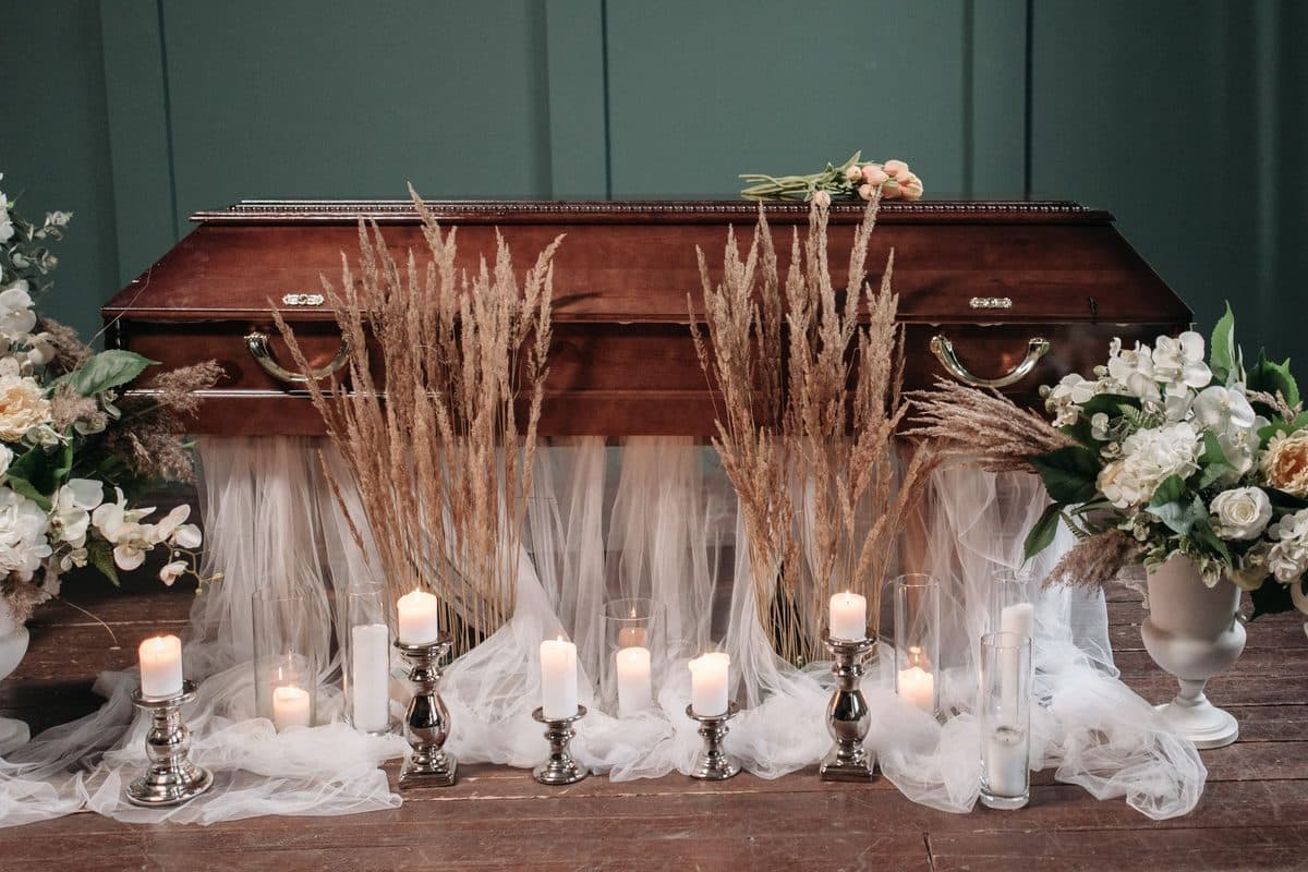 A wooden coffin in a funeral room with lit candles, white flowers and dried grasses in front of a green wall