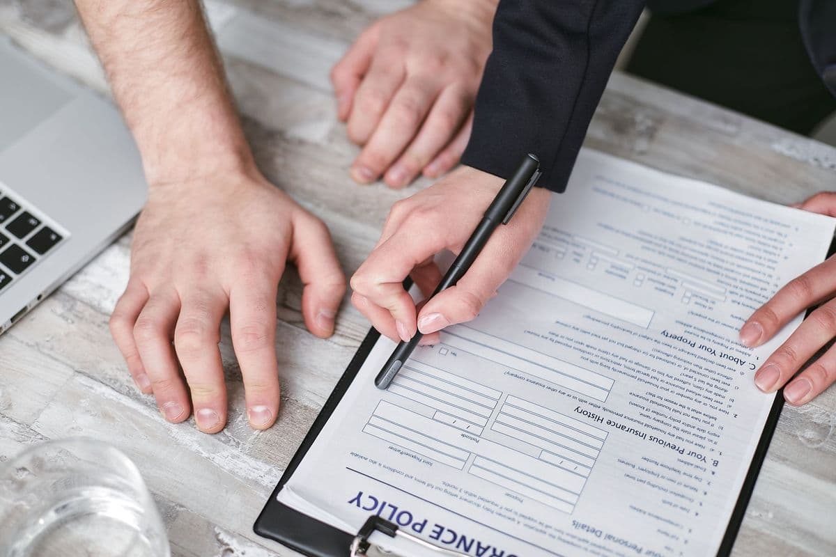 Two people's hands over an insurance policy application on a clipboard, one holding a pen, beside a laptop on a wooden desk