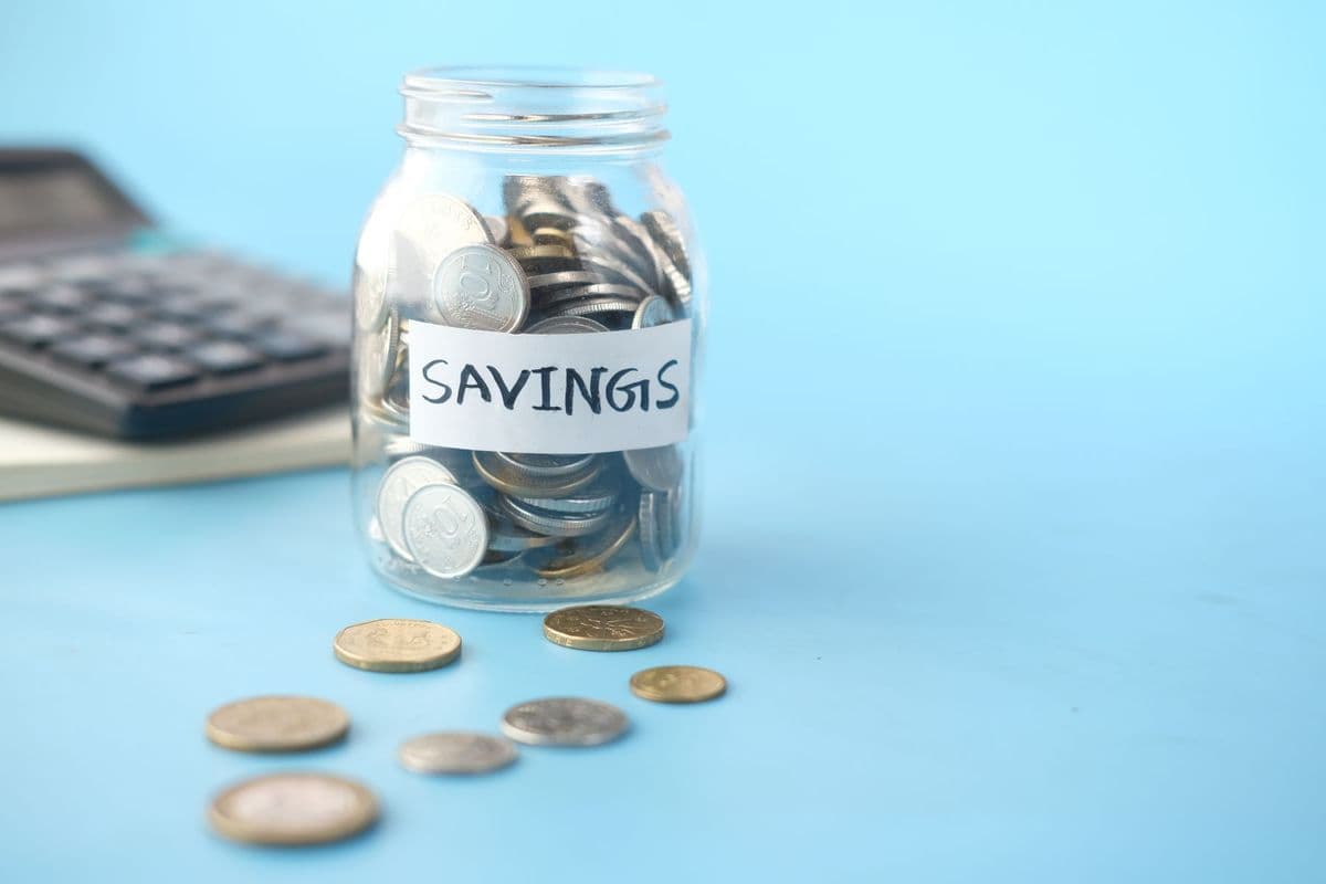 A glass jar of coins labelled Savings on a blue table, with loose coins in front and a calculator behind