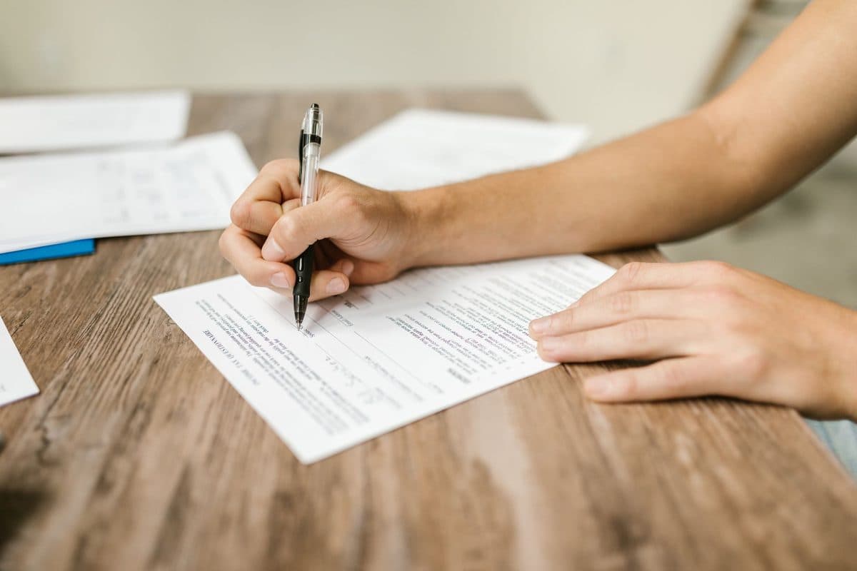 A hand filling out a printed form with a pen on a wooden desk