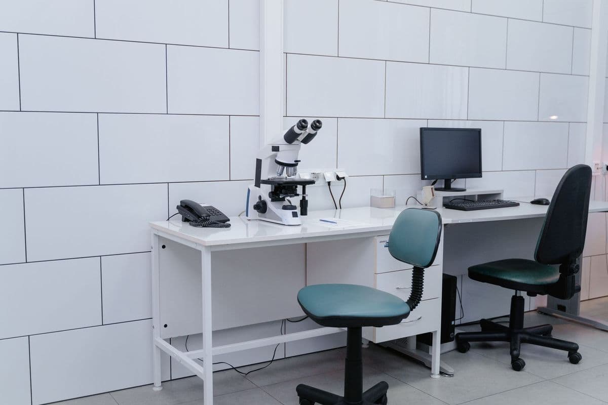 An empty white laboratory with a microscope, a desk phone and a computer monitor on white desks, and two teal office chairs
